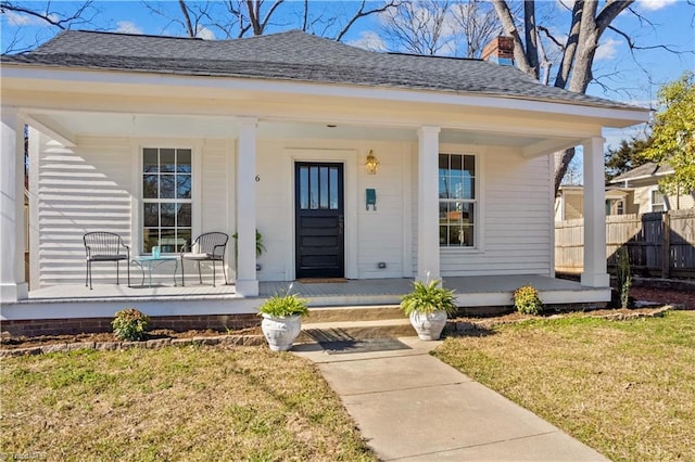 entrance to property with roof with shingles, a chimney, covered porch, a lawn, and fence