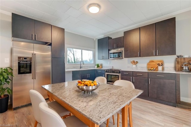 kitchen featuring appliances with stainless steel finishes, light stone counters, dark brown cabinetry, sink, and light hardwood / wood-style flooring