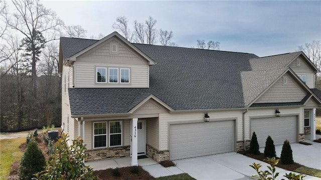 craftsman-style home featuring cooling unit, driveway, an attached garage, a shingled roof, and stone siding