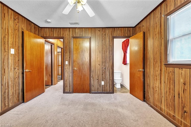 unfurnished bedroom featuring wood walls, ensuite bath, a textured ceiling, light carpet, and ceiling fan