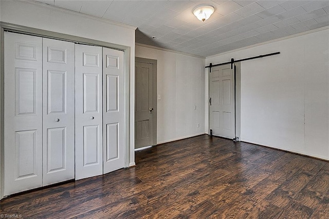 unfurnished bedroom featuring a barn door, baseboards, dark wood-style flooring, crown molding, and a closet