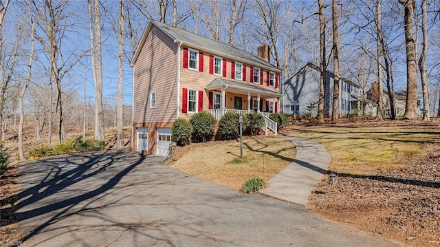 colonial inspired home with a porch, aphalt driveway, an attached garage, a chimney, and a front yard