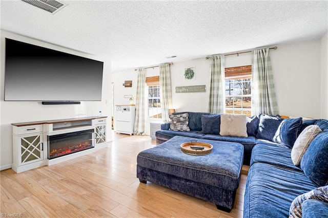 living room with a textured ceiling, light wood-style flooring, a glass covered fireplace, and visible vents