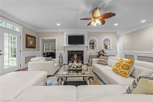 living room featuring a glass covered fireplace, ornamental molding, a ceiling fan, wood finished floors, and a wainscoted wall