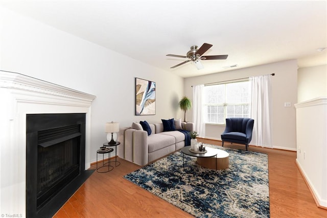 living room featuring baseboards, visible vents, a fireplace with flush hearth, ceiling fan, and wood finished floors