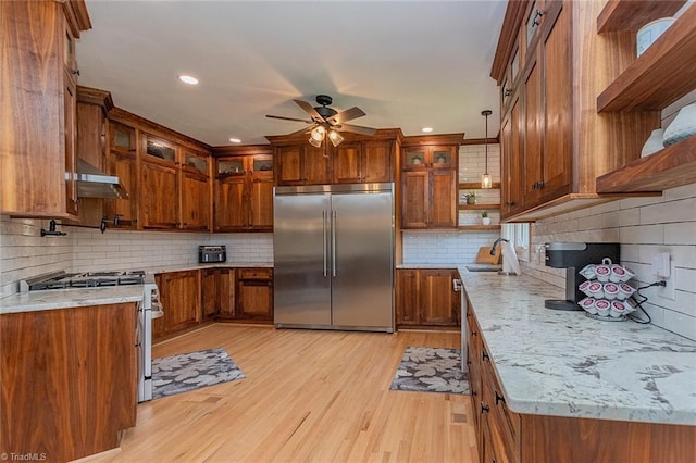 kitchen featuring light stone counters, hanging light fixtures, stainless steel appliances, range hood, and light hardwood / wood-style floors