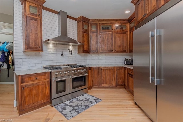 kitchen featuring tasteful backsplash, premium appliances, light stone counters, wall chimney range hood, and light wood-type flooring