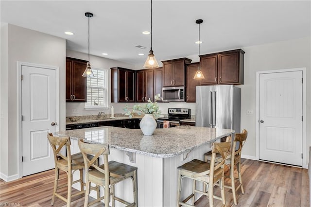kitchen featuring light wood-style flooring, stainless steel appliances, dark brown cabinetry, a kitchen island, and light stone counters