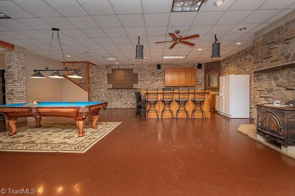 playroom with ceiling fan, brick wall, a drop ceiling, and a wood stove