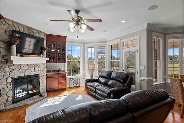 living room featuring ornamental molding, light wood-type flooring, a stone fireplace, and a ceiling fan