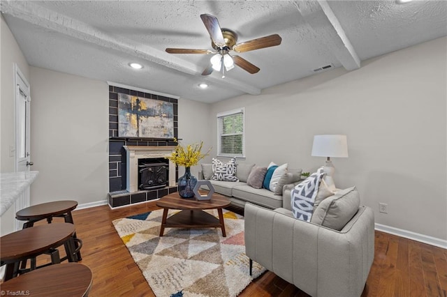 living room featuring a textured ceiling, ceiling fan, hardwood / wood-style flooring, beamed ceiling, and a tiled fireplace