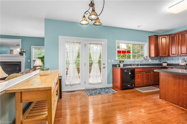 kitchen featuring black dishwasher, light wood-type flooring, backsplash, a glass covered fireplace, and decorative light fixtures