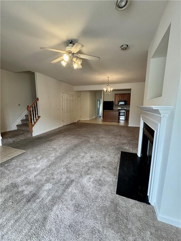 unfurnished living room with carpet, a ceiling fan, a glass covered fireplace, stairway, and a chandelier