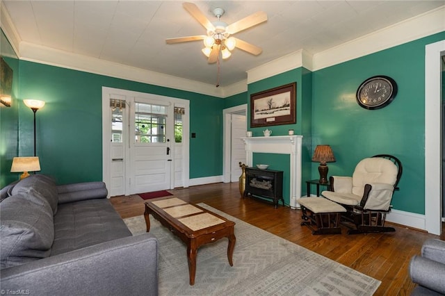 living room with crown molding, wood finished floors, a ceiling fan, baseboards, and a wood stove