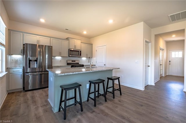 kitchen featuring appliances with stainless steel finishes, gray cabinets, a center island with sink, a breakfast bar, and dark wood-style floors