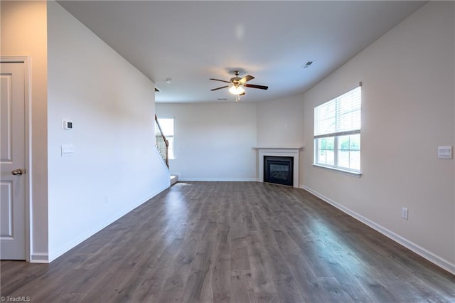 unfurnished living room featuring a ceiling fan, dark wood finished floors, baseboards, a glass covered fireplace, and stairs