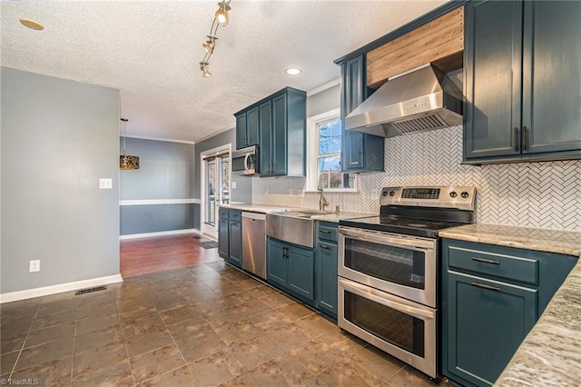 kitchen with pendant lighting, wall chimney range hood, appliances with stainless steel finishes, backsplash, and light stone counters