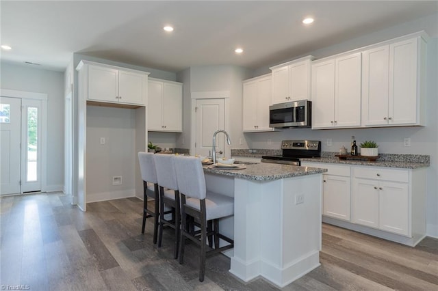 kitchen with appliances with stainless steel finishes, wood finished floors, a kitchen island with sink, white cabinets, and recessed lighting