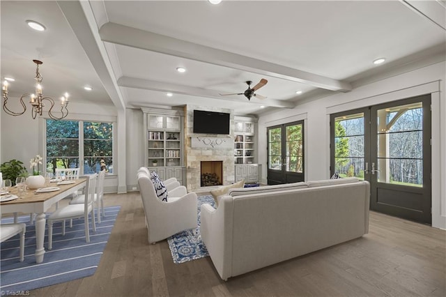 living room featuring french doors, a large fireplace, hardwood / wood-style flooring, beam ceiling, and ceiling fan with notable chandelier