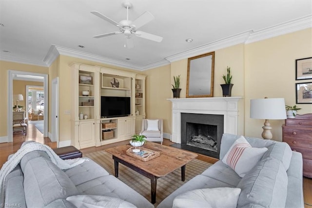 living room featuring a ceiling fan, wood finished floors, baseboards, a fireplace with flush hearth, and ornamental molding