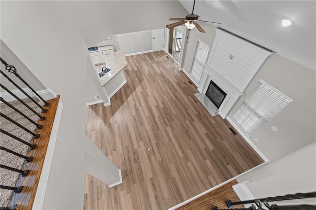unfurnished living room with ceiling fan, light wood finished floors, a fireplace with raised hearth, stairway, and baseboards