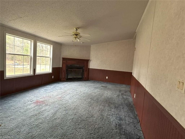 unfurnished living room featuring a ceiling fan, carpet, a wainscoted wall, a fireplace, and a textured ceiling