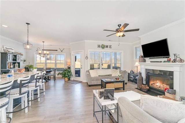 living area with ceiling fan with notable chandelier, wood finished floors, a glass covered fireplace, and crown molding