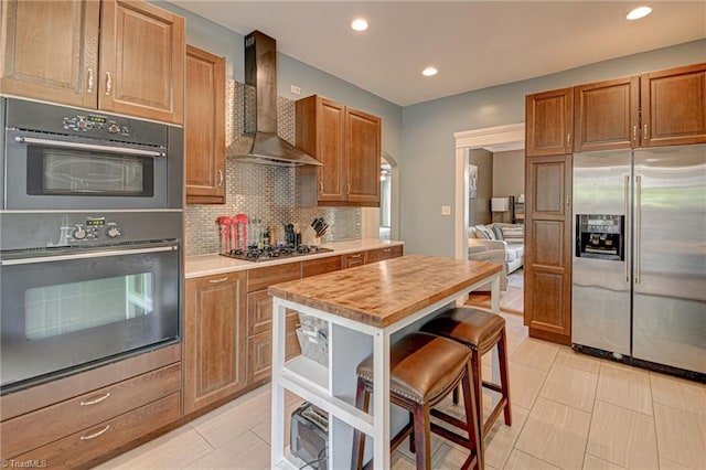 kitchen with stainless steel appliances, wall chimney range hood, tasteful backsplash, recessed lighting, and brown cabinetry