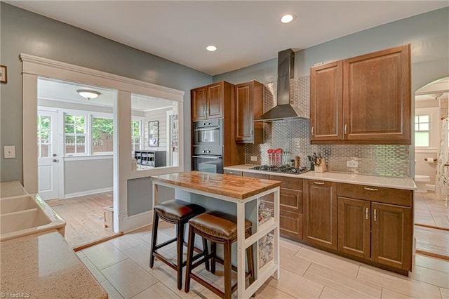kitchen with wall chimney exhaust hood, double oven, stainless steel gas cooktop, arched walkways, and tasteful backsplash