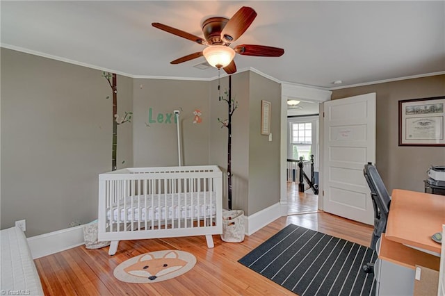 bedroom with wood finished floors, crown molding, baseboards, ceiling fan, and a crib
