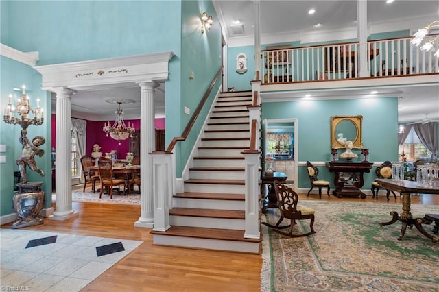 stairway with a towering ceiling, ornate columns, wood-type flooring, ornamental molding, and a notable chandelier