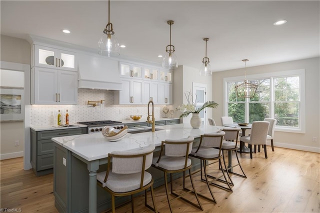 kitchen featuring a sink, custom exhaust hood, backsplash, light wood finished floors, and a center island with sink