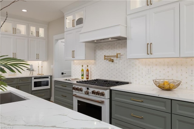 kitchen featuring white gas range oven, custom exhaust hood, light stone counters, white cabinets, and decorative backsplash