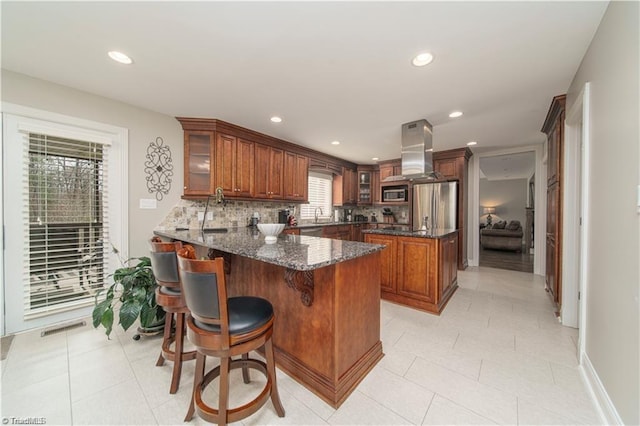 kitchen featuring island range hood, appliances with stainless steel finishes, a peninsula, dark stone countertops, and brown cabinetry