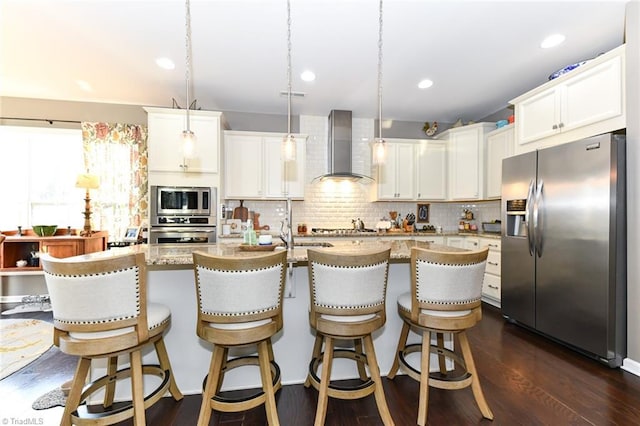 kitchen with an island with sink, white cabinets, hanging light fixtures, stainless steel appliances, and wall chimney exhaust hood