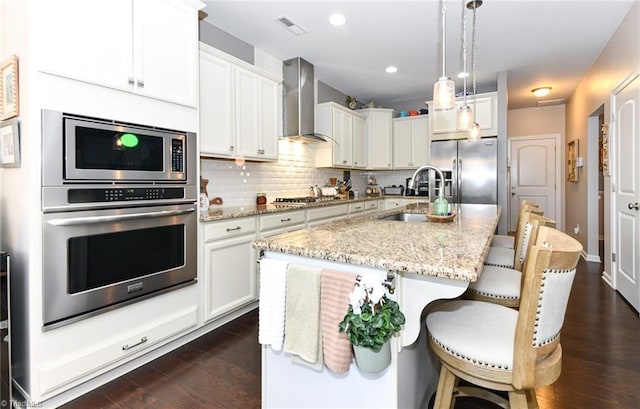 kitchen featuring wall chimney exhaust hood, white cabinetry, appliances with stainless steel finishes, pendant lighting, and a kitchen island with sink