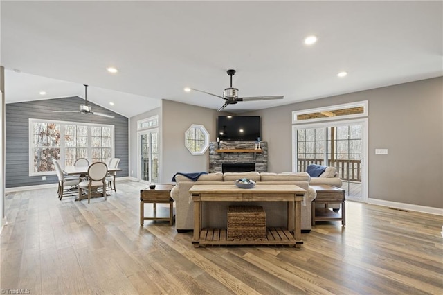 living area featuring a ceiling fan, vaulted ceiling, a stone fireplace, and light wood finished floors