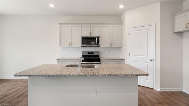 kitchen featuring appliances with stainless steel finishes, dark hardwood / wood-style flooring, light stone counters, and an island with sink