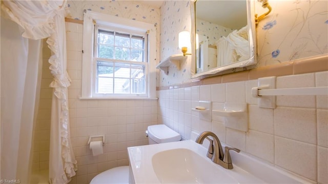 bathroom featuring wallpapered walls, toilet, tile walls, a sink, and a wainscoted wall