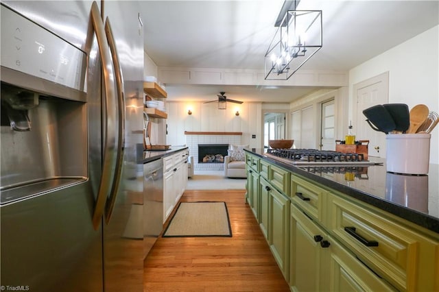 kitchen with light wood finished floors, ceiling fan, stainless steel appliances, green cabinets, and a brick fireplace