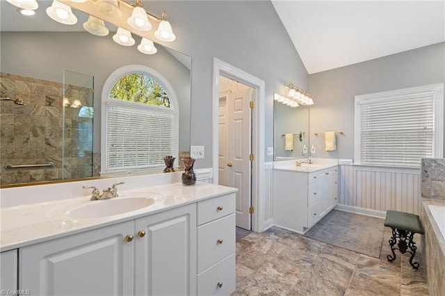 full bath featuring a wainscoted wall, tiled shower, lofted ceiling, two vanities, and a sink