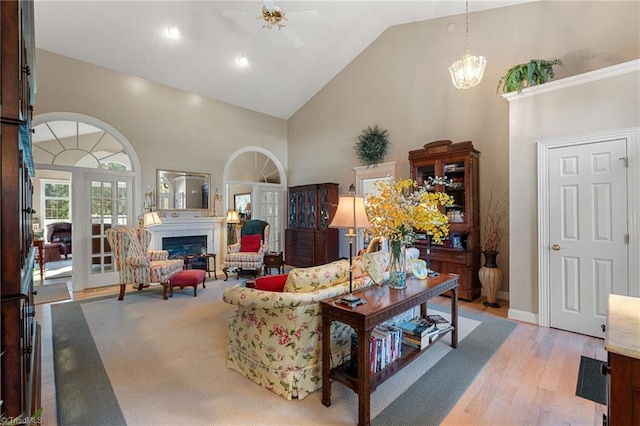 living area with baseboards, ceiling fan with notable chandelier, light wood-style floors, a glass covered fireplace, and high vaulted ceiling