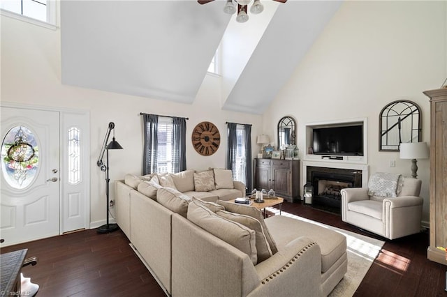 living area featuring baseboards, ceiling fan, dark wood-style floors, a glass covered fireplace, and high vaulted ceiling