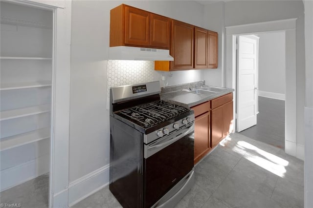 kitchen featuring under cabinet range hood, a sink, stainless steel gas range, brown cabinets, and tasteful backsplash