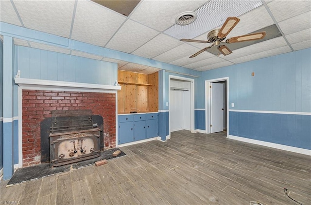 unfurnished living room featuring a wood stove, ceiling fan, a drop ceiling, and dark hardwood / wood-style floors