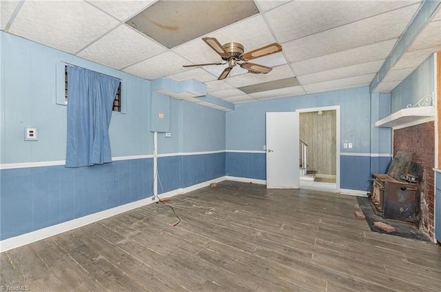 unfurnished living room featuring hardwood / wood-style flooring, a drop ceiling, ceiling fan, and a wood stove