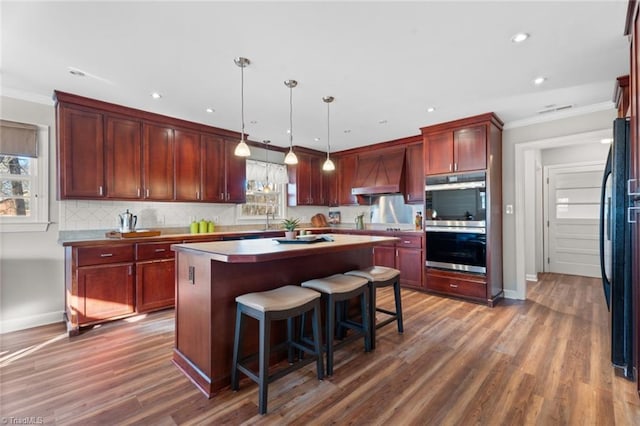 kitchen with black refrigerator, premium range hood, dark wood-type flooring, a kitchen island, and hanging light fixtures