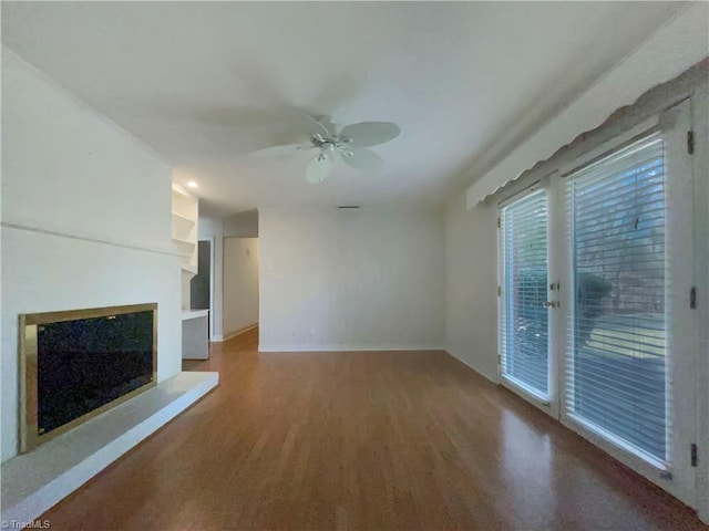 unfurnished living room with built in shelves, a fireplace with raised hearth, ceiling fan, and wood finished floors