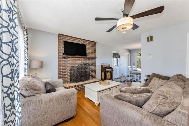 living room featuring a brick fireplace, a textured ceiling, light wood finished floors, and ceiling fan