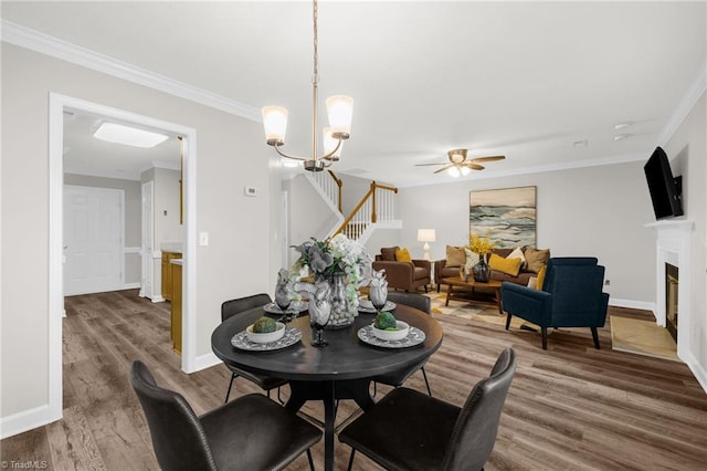 dining room featuring a ceiling fan, a fireplace, wood finished floors, ornamental molding, and stairs